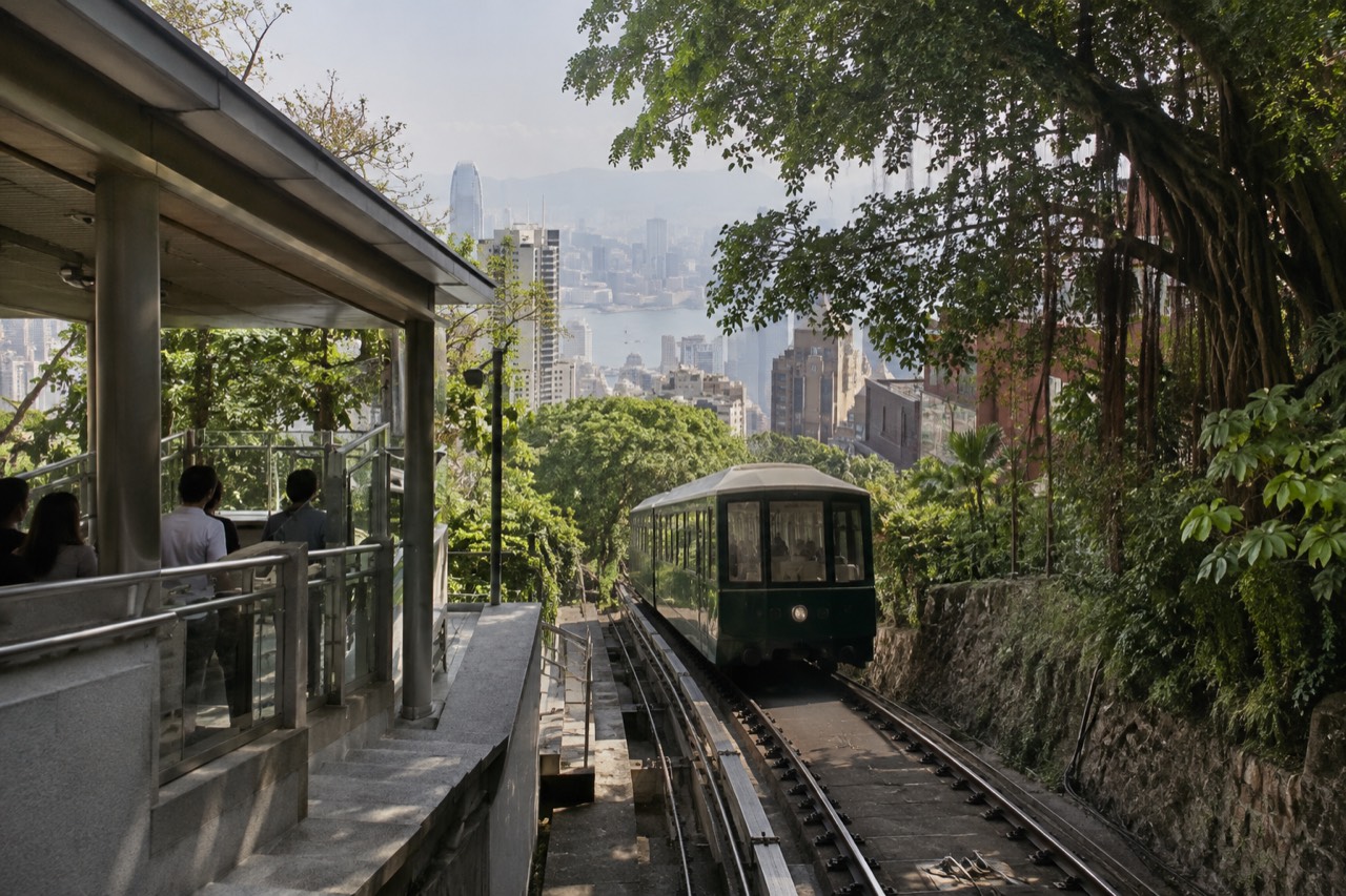 A green Peak Tram funicular car climbing a forested slope with Hong Kong skyscrapers below