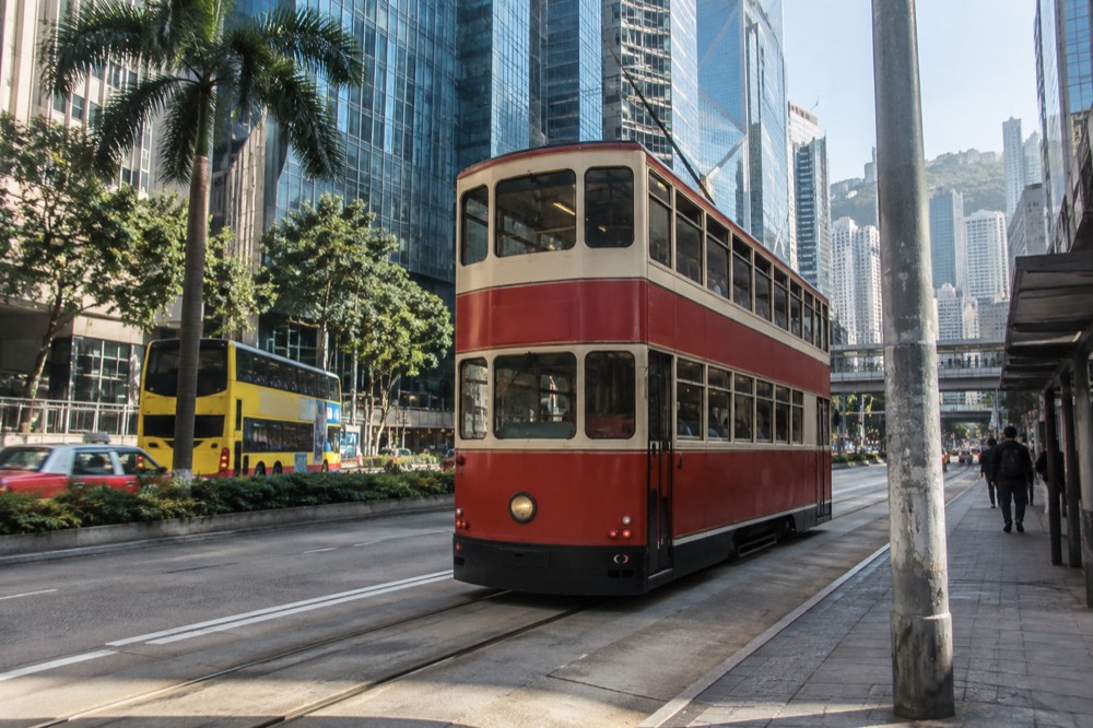 A red double-decker street tram running along a flat road beside Hong Kong skyscrapers