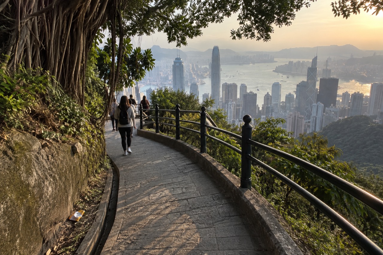 A paved hillside path curving downhill past trees toward Hong Kong harbour in the distance