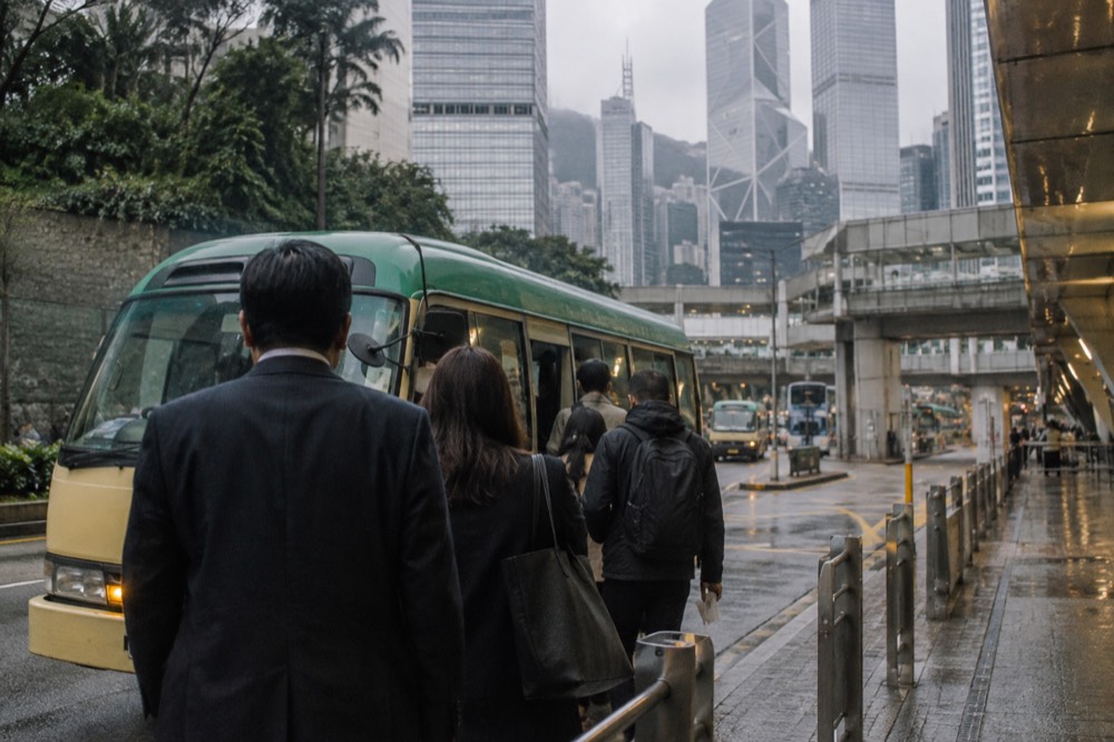 Passengers boarding a green minibus at a roadside stop beside tall office towers