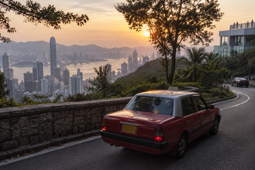 A red urban taxi on a curving mountain road with harbour views behind