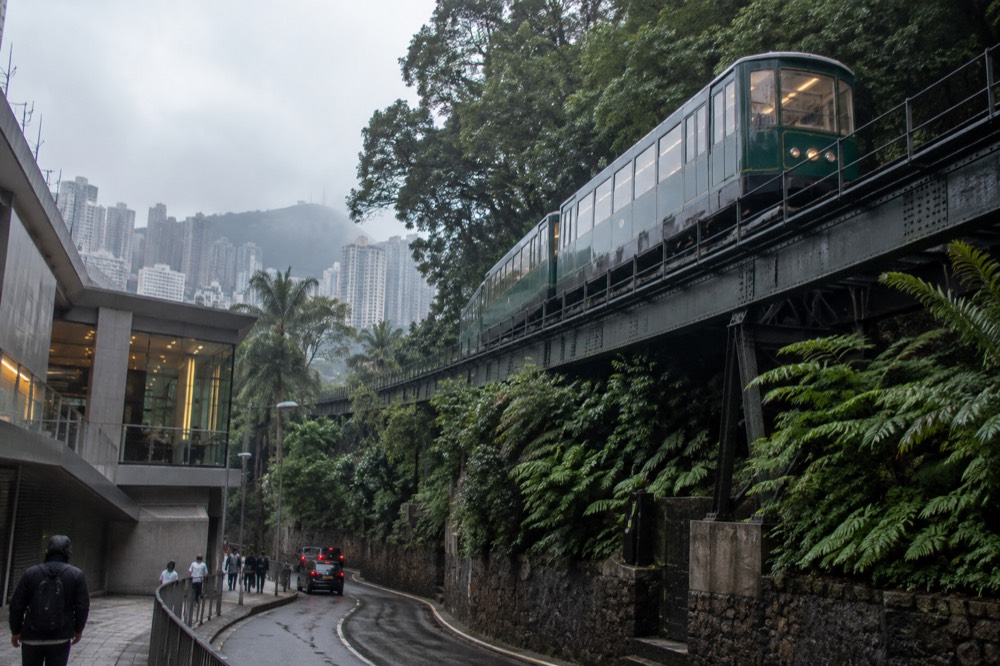 A green funicular tramcar on elevated track above a curved road on a forested hillside